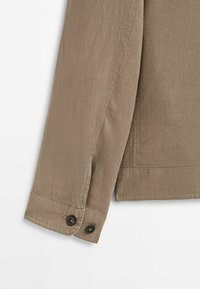 Manche d'une veste en lin beige avec deux boutons noirs sur le poignet et des coutures visibles sur un fond blanc.