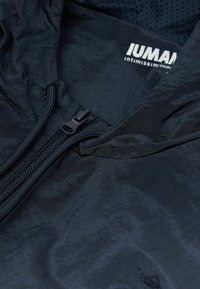 Lättvikts svart zip-up jacka med en texturerad yta. Inkluderar ett meshfoder och en frontdragkedja, med varumärket synligt vid halsringningen.