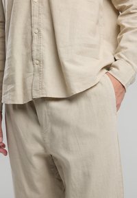 Vaaleanbeige pellavapaita, jossa on rento istuvuus. Paitassa on napitettava etuosa, pitkät hihat ja sivutasku, ja se on yhdistetty istuviin vaaleanbeigeihin housuihin.