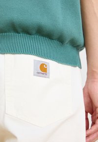 Maglione lavorato a maglia verde menta con un orlo a coste, abbinato a pantaloni color crema con una toppa rettangolare grigia del logo Carhartt sulla tasca posteriore.