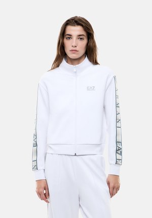Donna che indossa una giacca bianca con zip e pantaloni abbinati con il marchio "EA7 Emporio Armani" sulle maniche e sul petto su uno sfondo semplice.