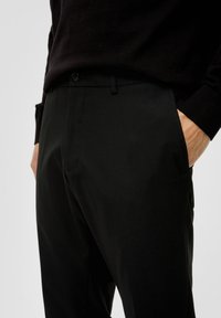 Zwarte op maat gemaakte broek van gladde stof, met een slim fit, riemlussen en zijzakken. Knopsluiting bij de taille zichtbaar.