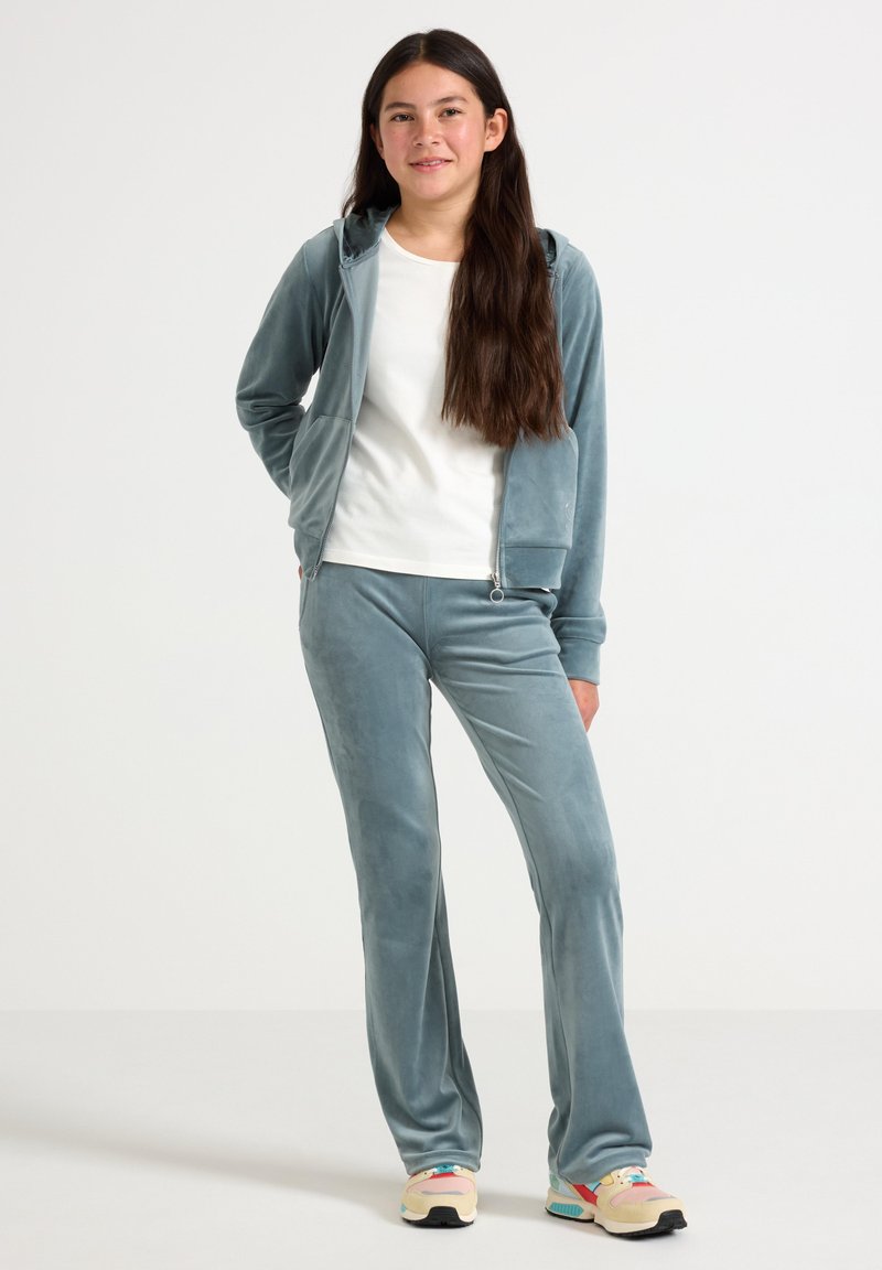 Velvet tracksuit in soft blue, featuring a zip-up hoodie and matching pants with a smooth texture. White T-shirt underneath.
