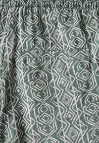 Tela verde con un patrón geométrico blanco que presenta diamantes y bucles; incluye un bolsillo trasero y una cintura elástica.