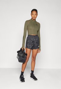 Haut à manches longues cropped couleur olive, shorts noirs à imprimé animal, chaussures de sport noires et un sac de sport noir avec un logo blanc.