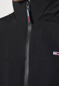 Černá bunda s vysokým límcem a předním voděodolným zipem, který má poutko v modré, bílé a červené barvě.