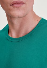 Green cotton t-shirt with a round neckline. Smooth texture with subtle ribbing on the collar. Close-up view of the neck and shoulders.