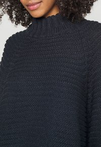 Marinblått stickat tröja med hög ribbad krage, med en strukturerad mönster och lös passform. Tyget verkar mjukt och varmt.