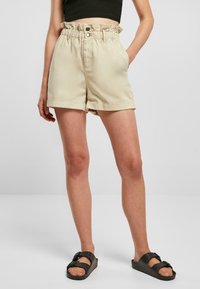 Beige høytlivet shorts laget av mykt stoff, med volang i livet, to sidelommer og oppbrettede kanter. Brukt med svarte sandaler.