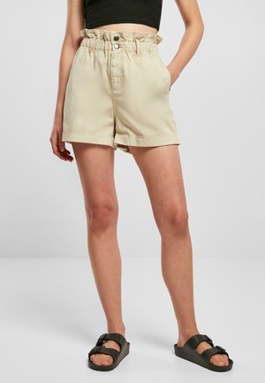Shorts beiges taille haute en tissu doux, avec une taille froncée, deux poches latérales et des ourlets retroussés. Portés avec des sandales noires.