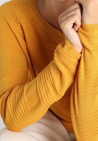 Pull en laine jaune tricoté avec une texture côtelée et une légère variation du motif sur les manches. Gros plan sur les bras et les mains.