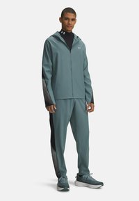Teal sportjack met een ritssluiting, zwarte accenten aan de zijkanten en witte strepen; bijpassende broek; ademend, lichtgewicht materiaal; trainingsschoenen.