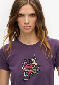 Camiseta de algodón púrpura con un diseño bordado que incluye un corazón, flores y una cinta que dice "POR SIEMPRE." Manga corta y cuello redondo.