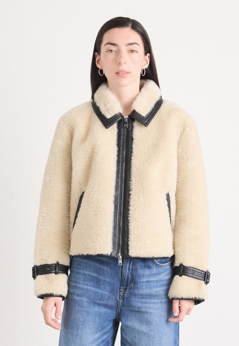 Beige nepleerjack met zwarte leren accenten, ritssluiting aan de voorkant en brede kraag. Beschikt over verstelbare manchetten met gespdetails.