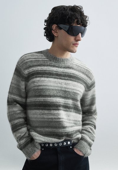 Pull rayé aux teintes douces de gris et crème, col rond et coupe décontractée, associé à une ceinture noire ornée de clous métalliques.