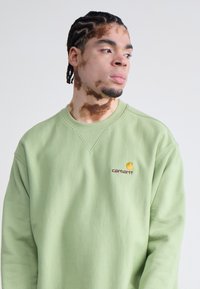 Lys grøn sweatshirt lavet af blødt materiale, med rund hals og et lille broderet logo i gul og burdgund på brystet.