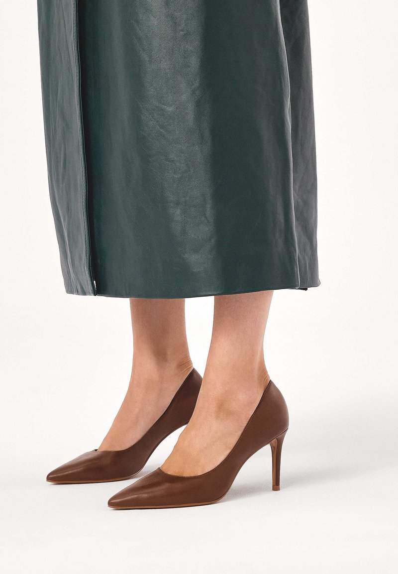 Donna che indossa décolleté marroni a punta abbinati a una gonna midi in pelle verde scuro, in piedi su un pavimento bianco.