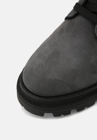 Chaussure en cuir gris avec une surface texturée, un bout rond et une semelle extérieure en caoutchouc noir présentant des motifs de crampons proéminents. Détail de laçage visible.