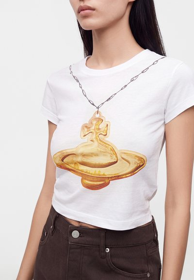 Γυναίκα που φοράει λευκό crop t-shirt με μεγάλο χρυσό μενταγιόν και σχέδιο αλυσίδας, σε συνδυασμό με σκούρο τζιν ψηλόμεσο.