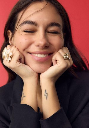 Junge Frau lächelt mit geschlossenen Augen, Hände ruhen am Kinn, trägt Blumen- und Edelsteinringe, sichtbare Tätowierungen eines Sterns und eines Blitzes am Handgelenk.