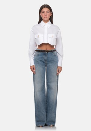 Donna che indossa una camicia bianca corta con bottoni e due tasche frontali e jeans blu a vita alta a gamba larga con cintura nera.