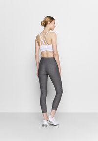 Vit sport-bh med korsad ryggdesign, i kombination med åtsittande grå leggings. Modellen bär vita träningsskor. Minimalistisk bakgrund.