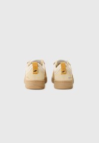 Beige suede sneakers with a tan rubber sole. Features a yellow pull tab at the heel and minimal branding on the back.