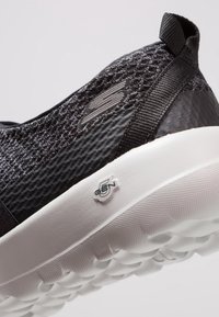 Chaussure de sport tricotée noire avec une tige texturée, semelle en caoutchouc flexible et logo blanc proéminent sur le talon ; accents lisses.