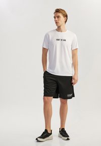 Jonge man die staat te poseren in een witte "FIGHT TO WIN" t-shirt, zwarte shorts met een tekstpatch, en zwart-witte sneakers tegen een effen achtergrond.
