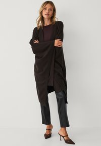 Mørkebrun cardigan lavet af blødt stof med brede ærmer, lagdelt over en maroon top, paired med sorte fake læderbukser og brune hæle.