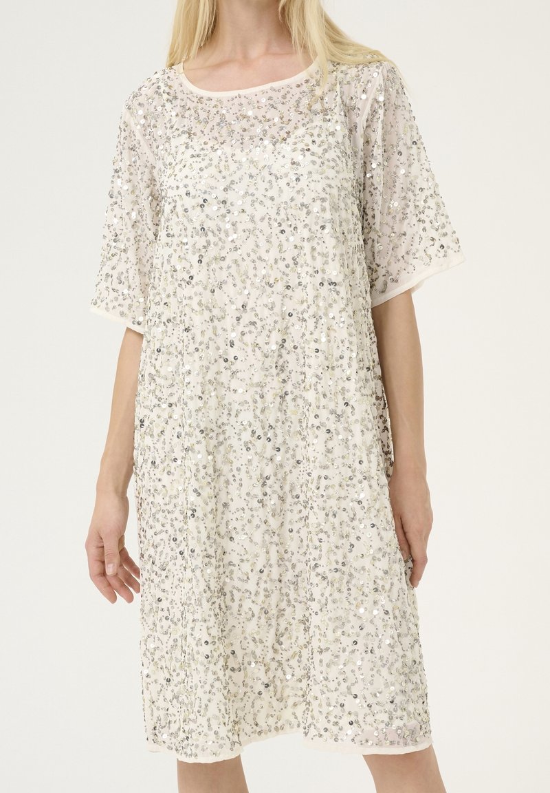 Femme portant une robe ample mi-longue à manches courtes, couverte de sequins argentés sur un tissu transparent pâle.