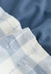 Nærbillede af blåt stof med en foldet lyseblå og hvidternet tekstilkant, der viser detaljeret syning og tekstur.