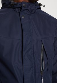 Navy waterproof jacket featuring a high collar, zippered chest pocket, and textured fabric with visible stitching details.