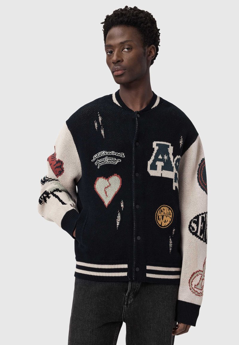 Giovane uomo che indossa una giacca varsity blu navy e beige con toppe, tra cui un cuore spezzato, lettere e vari simboli, in piedi davanti a uno sfondo semplice.