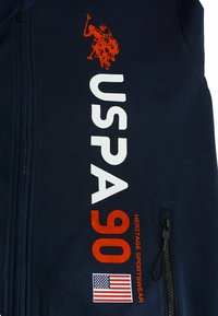 Férfi sötétkék cipzáras dzseki narancssárga póló játékos grafikával, feltűnő fehér "USPA" és "90" felirattal, valamint amerikai zászló részlettel. Simogatós textúra.