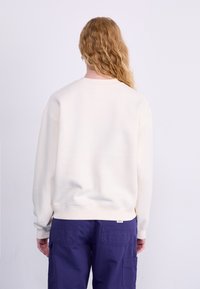 Sweatshirt couleur crème avec un col rond, des poignets et un ourlet côtelés. Porté avec un pantalon cargo violet foncé. Texture douce, design décontracté.