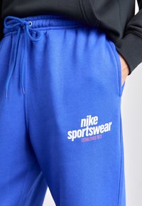 Detailní záběr osoby v modrých sportovních teplácích Nike s tkaničkou, ruka v kapse a viditelný rukáv černé mikiny.