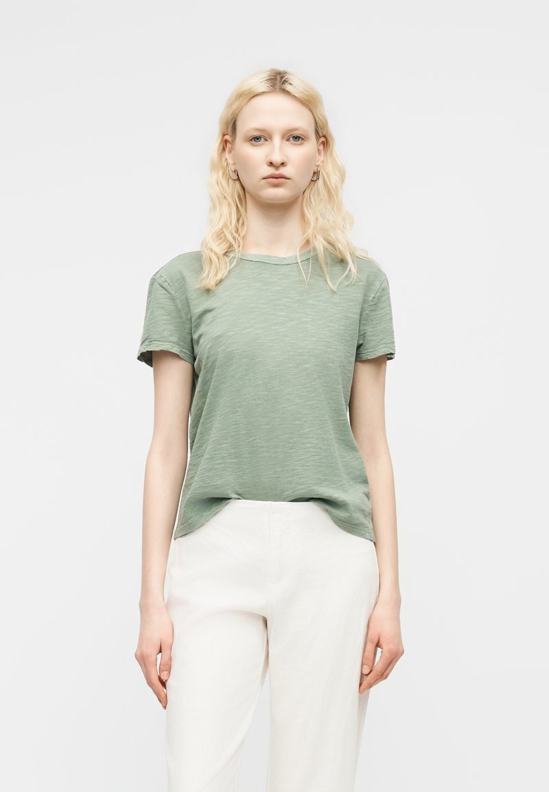Femme blonde portant une chemise à manches courtes vert clair et un pantalon blanc, debout devant un fond gris clair uni.