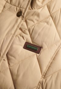 Veste matelassée beige avec un logo Superdry® marron bien visible ; présente des motifs en losange, une texture lisse et des détails de boutons métalliques.