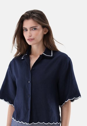 Frau mit langen braunen Haaren, die ein marineblaues Hemd mit Knöpfen trägt, mit wellenförmigen Kanten an Ärmeln und Saum, vor einem weißen Hintergrund.