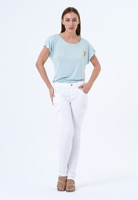 Haut bleu clair à manches courtes avec un détail brodé, associé à un pantalon cargo blanc avec poches latérales. Le modèle porte des sandales vertes à lanières.