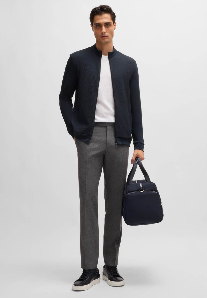 Veste zippée marine, t-shirt blanc, pantalon gris ajusté et baskets noires. Tenant un sac marine avec une fermeture éclair et des poignées courtes.