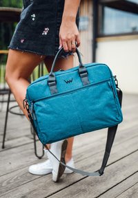 Türkisfarbene Laptop-Tasche aus Stoff mit zwei gepolsterten Griffen, verstellbarem grauen Tragegurt, vorderer Reißverschluss-Tasche und Logo-Akzent. Strukturierte Oberfläche, elegantes Design.