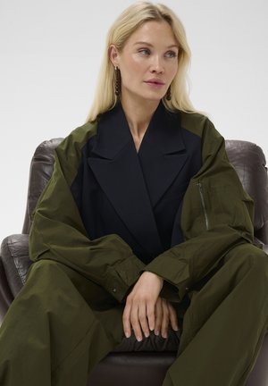 Blonde Frau in übergroßer grüner Jacke mit schwarzen Revers sitzt auf braunem Ledersessel, Hände gefaltet, schaut seitlich vor weißem Hintergrund.