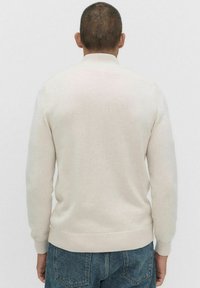 Jasnobeżowy sweter z prążkowanym kołnierzem i mankietami, dzianiną o fakturze oraz elastycznym dołem. Widoczny od tyłu, noszony z dżinsami.