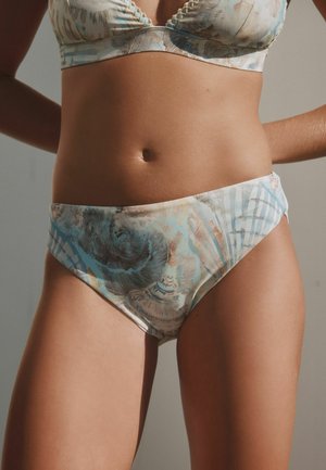 Frau, die pastellfarbene Bikinihosen mit Muschel-Muster und passendem Oberteil trägt, steht mit den Händen in der Hüfte vor einem einfarbigen Hintergrund.