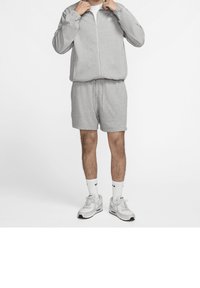 Chaqueta gris con cremallera y pantalones cortos a juego de tela suave, con un pequeño logo. Llevado con calcetines blancos y zapatillas, con acentos sutiles.