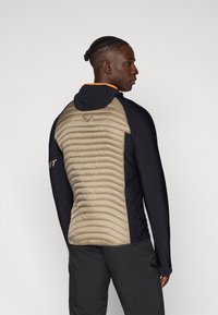 Veste isolante noire et beige avec un dos rembourré à texture côtelée, capuche et accents orange le long de l'encolure.