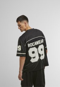 Fekete rövidujjú mez fehér csíkokkal. Nagy fehér "99" szám található a hátsó részen, felette pedig a "ROCAWEAR" felirat. Az anyag simának és légáteresztőnek tűnik.