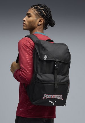 Jonge man met gevlochten haar die een rood Portugal-voetbalshirt draagt en een zwarte Puma Portugal-rugzak over één schouder draagt.